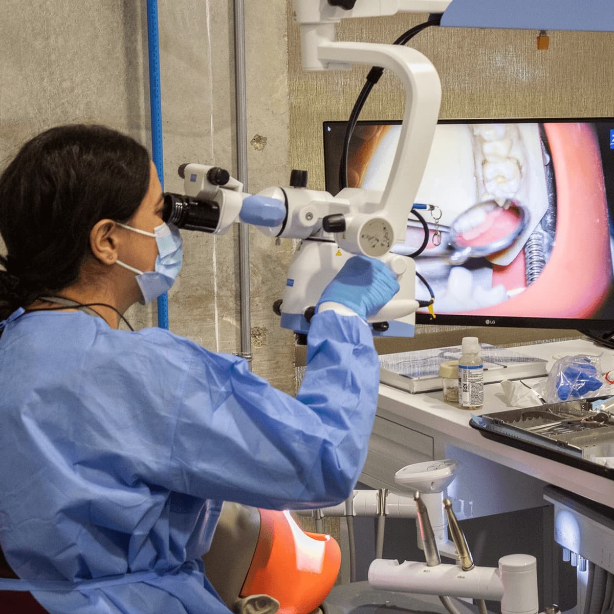 A participant at the microscope eyepieces with one hand on the scope body, adjusting it; the monitor behind shows a wide intraoral view of clamped molars