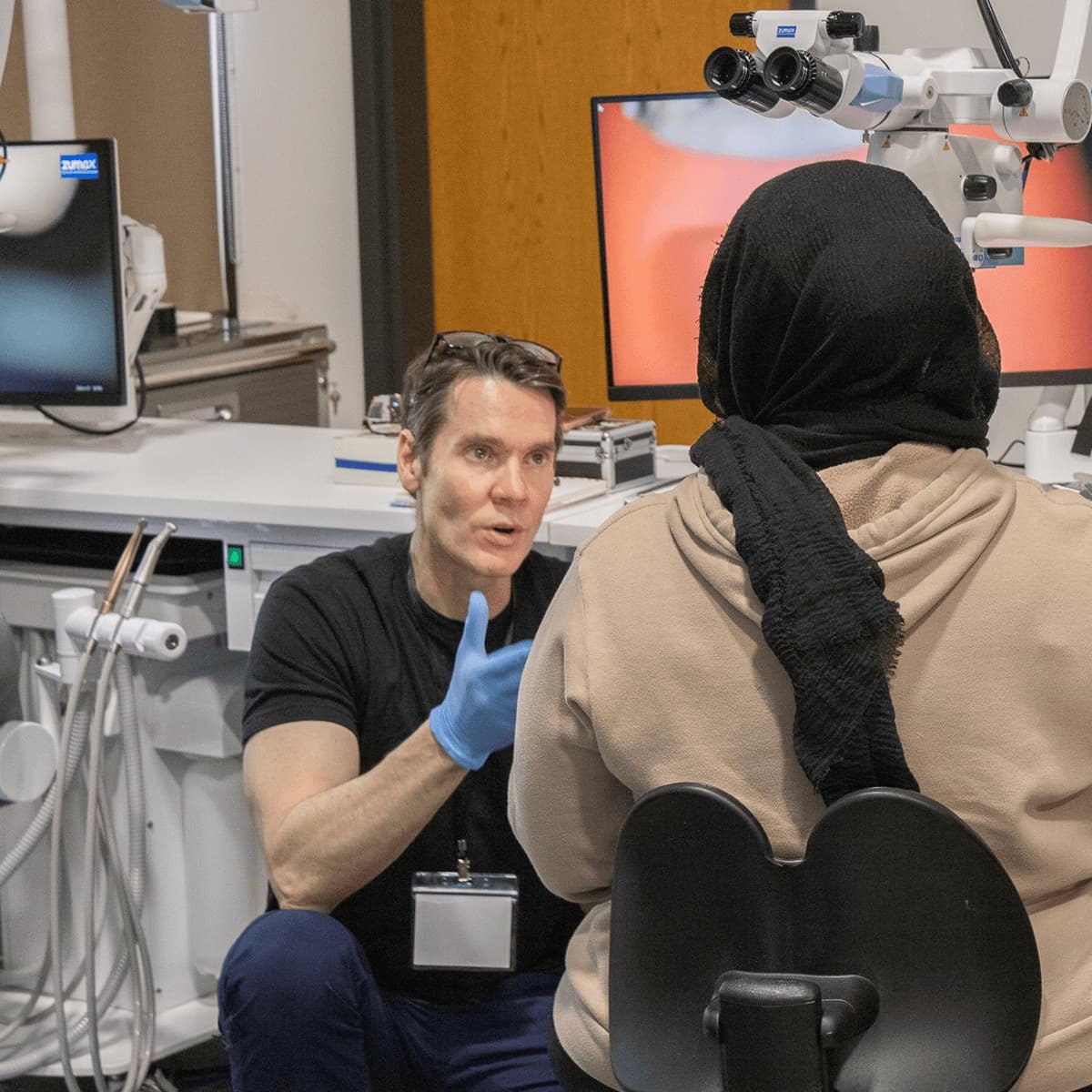 Dr. Nudera crouched beside a seated participant at her station, gloved hand raised mid-explanation as he talks her through a step