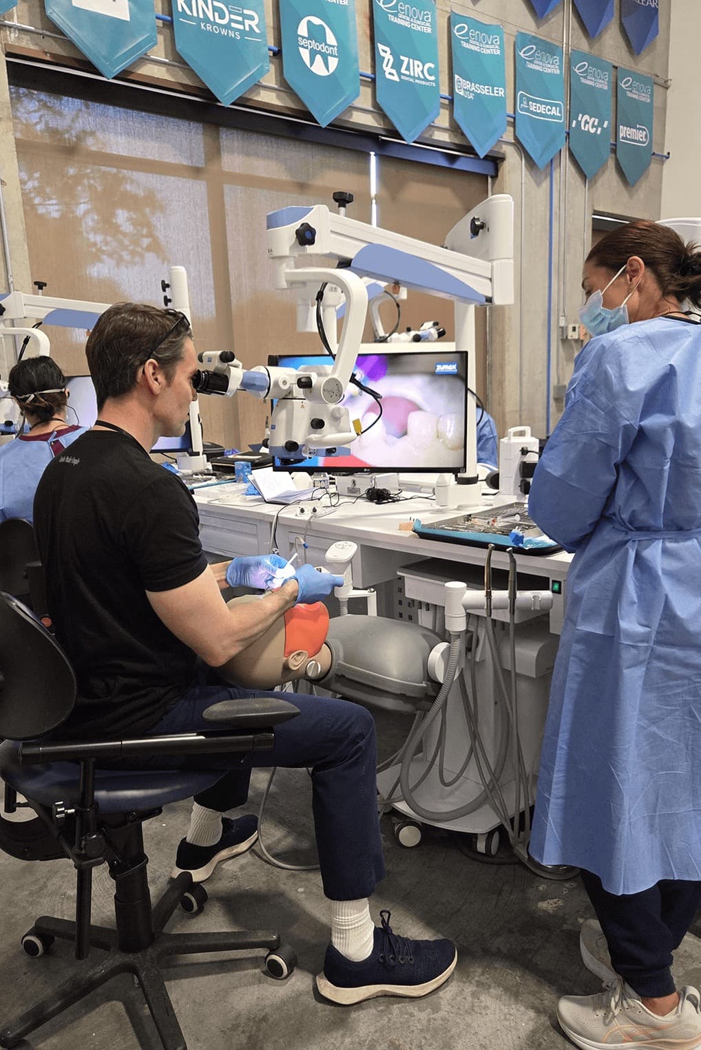A workshop participant treating a manikin tooth under a dental operating microscope, the magnified canal view on the monitor beside him, with an instructor observing at his shoulder